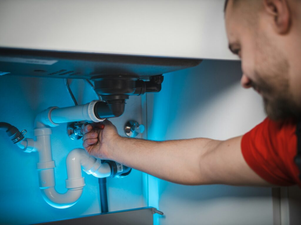 Plumber inspecting and working on pipes beneath a kitchen sink, assessing the plumbing system before a partial or whole home repipe.